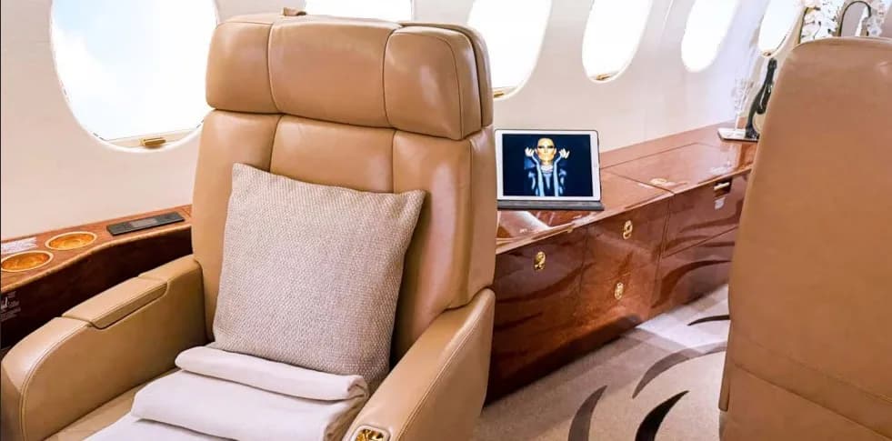 A close-up of a tan leather armchair in a private jet cabin, with a laptop displaying a person's face on a polished wooden console.