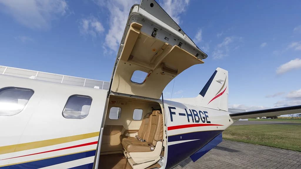The open passenger door of a white and blue TBM aircraft with registration F-HBGE, revealing the tan leather seats inside.