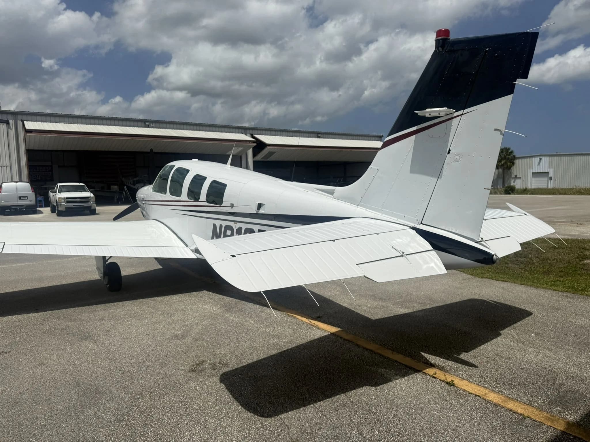 Rear three-quarter view of a white Beechcraft Bonanza N812BG with dark red and black stripes on an airfield.