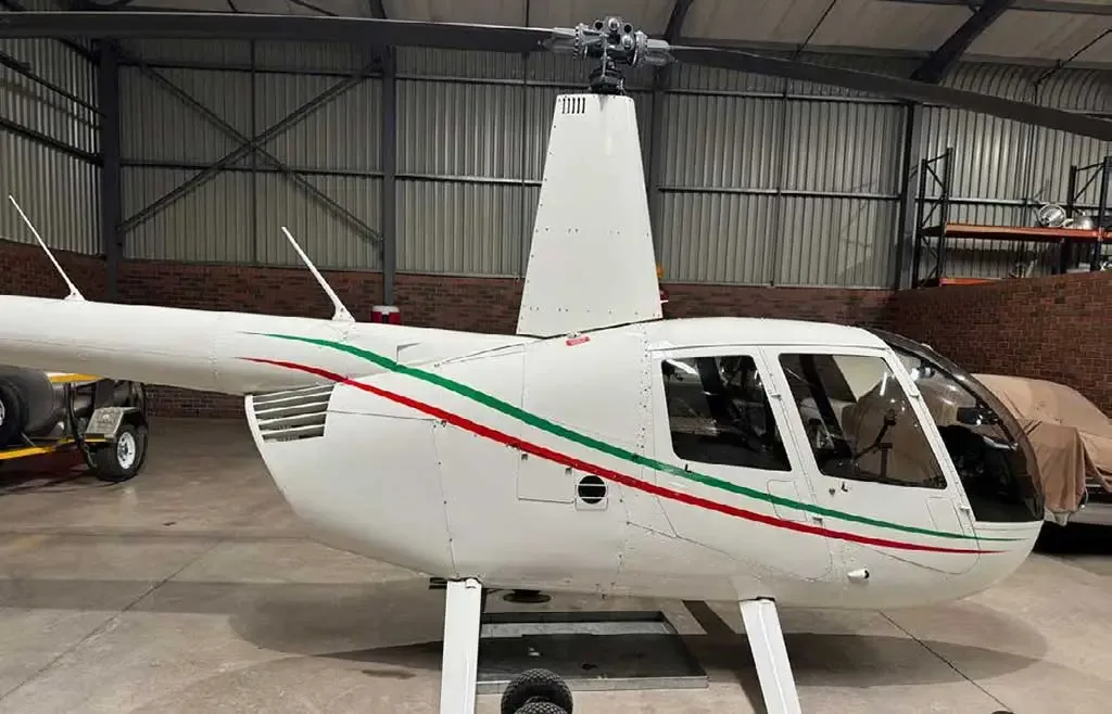 A white Robinson R44 helicopter with green and red stripes is parked inside a hangar, viewed from the right side.
