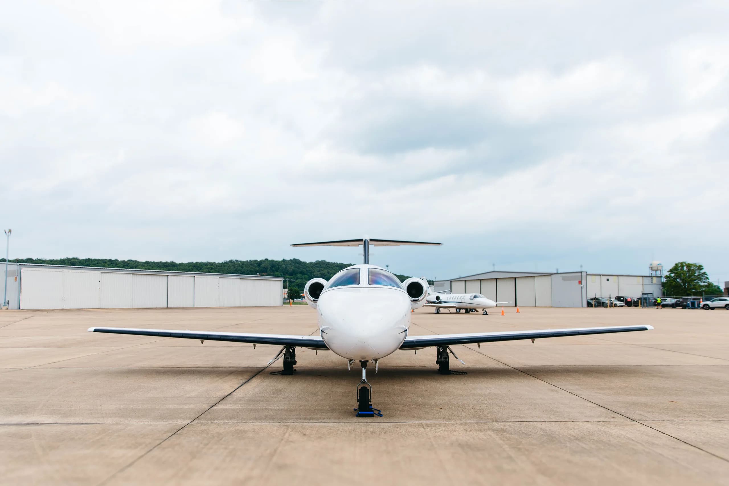 A frontal view of a white Cessna Citation M2 business jet, tail number N22EM, parked on a tarmac with another jet and hangars in the background.
