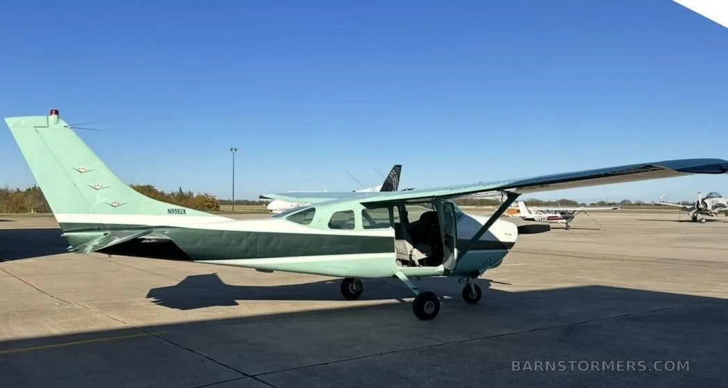 A light green and dark green Cessna 210 (N9982X) with its door open, parked on an airfield under a clear sky.