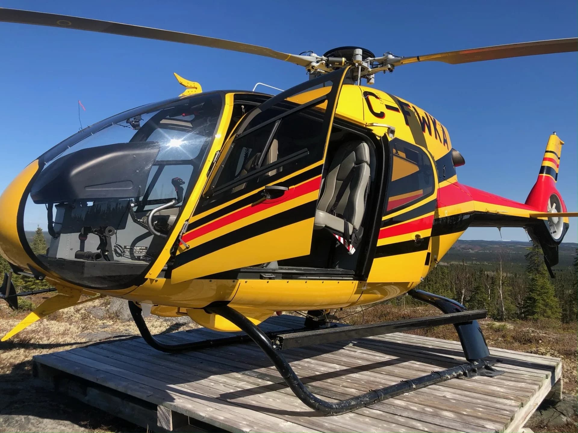 A yellow helicopter with red and black stripes, registration C-FWKA, parked on a wooden platform outdoors with its door open.
