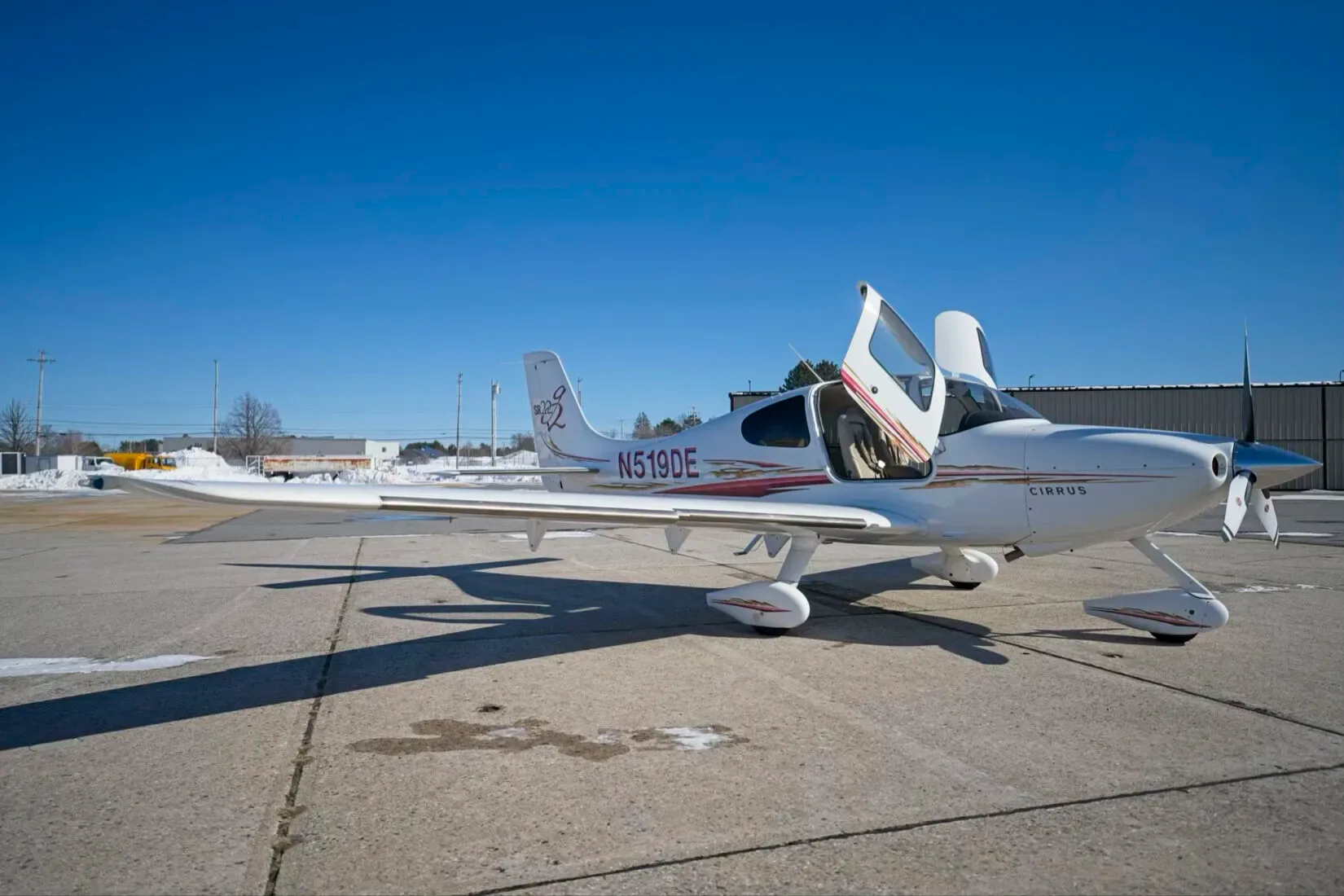 A white Cirrus SR22 aircraft with red and gold stripes, registration N519DE, parked on an airfield with its doors open.