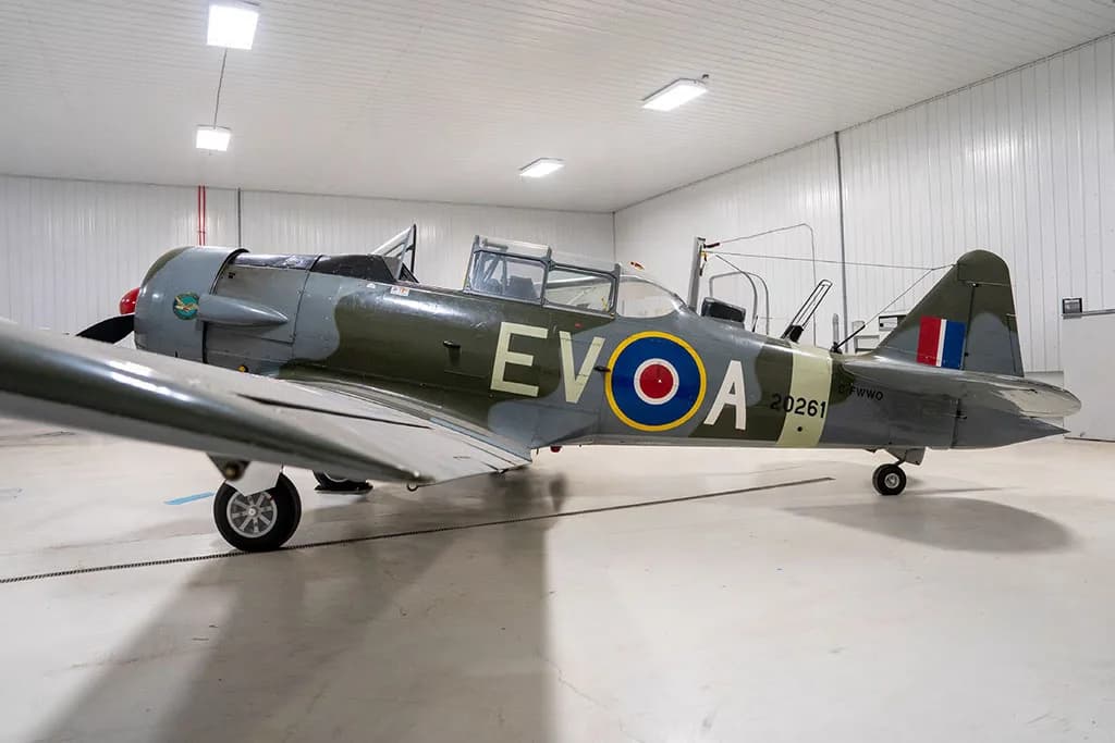 A side view of a North American Harvard Mk.IIB aircraft in green and grey camouflage with RAF roundels, parked in a hangar.