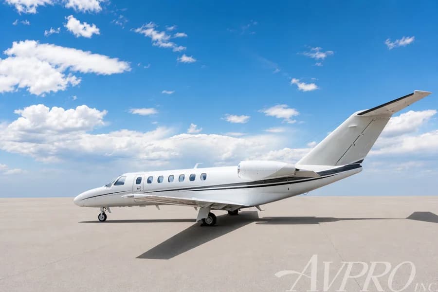 A white Cessna Citation jet with black and grey stripes parked on an airfield, viewed from the rear-left side.