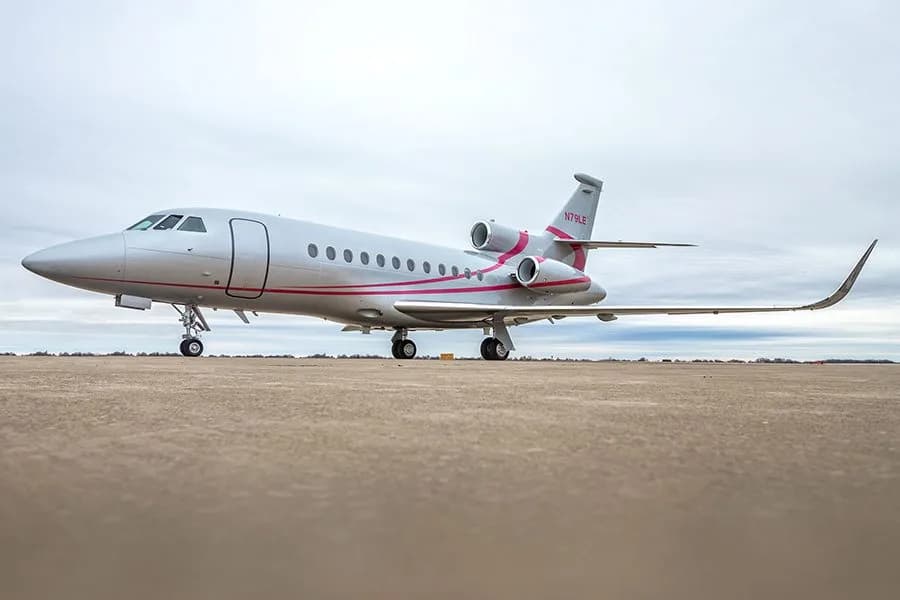 A low-angle side view of a silver Dassault Falcon 900EX private jet with pink accents on a concrete surface.