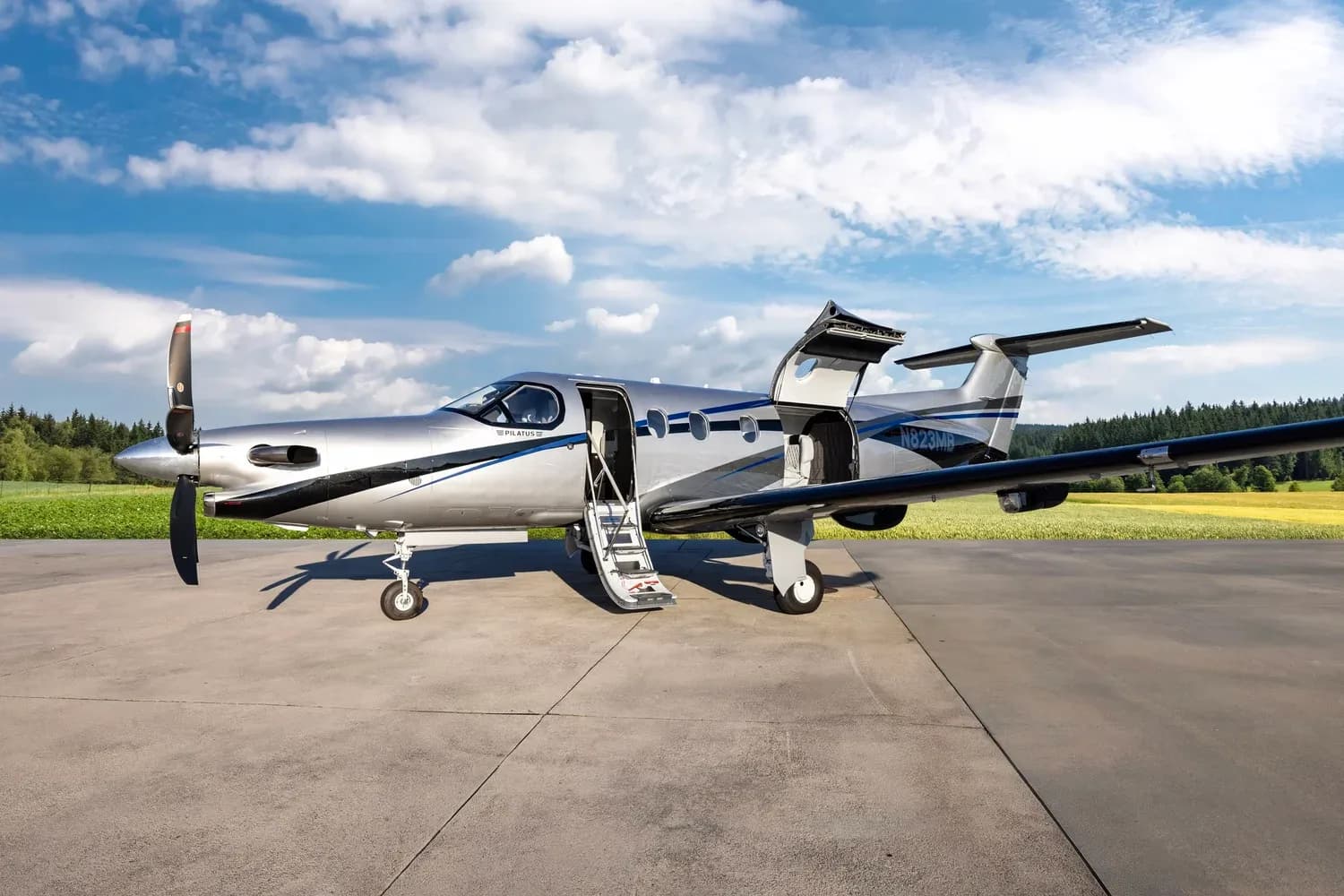 A silver Pilatus PC-12 NG aircraft, registration N823MB, with both its main passenger door and cargo door open, parked on a tarmac.