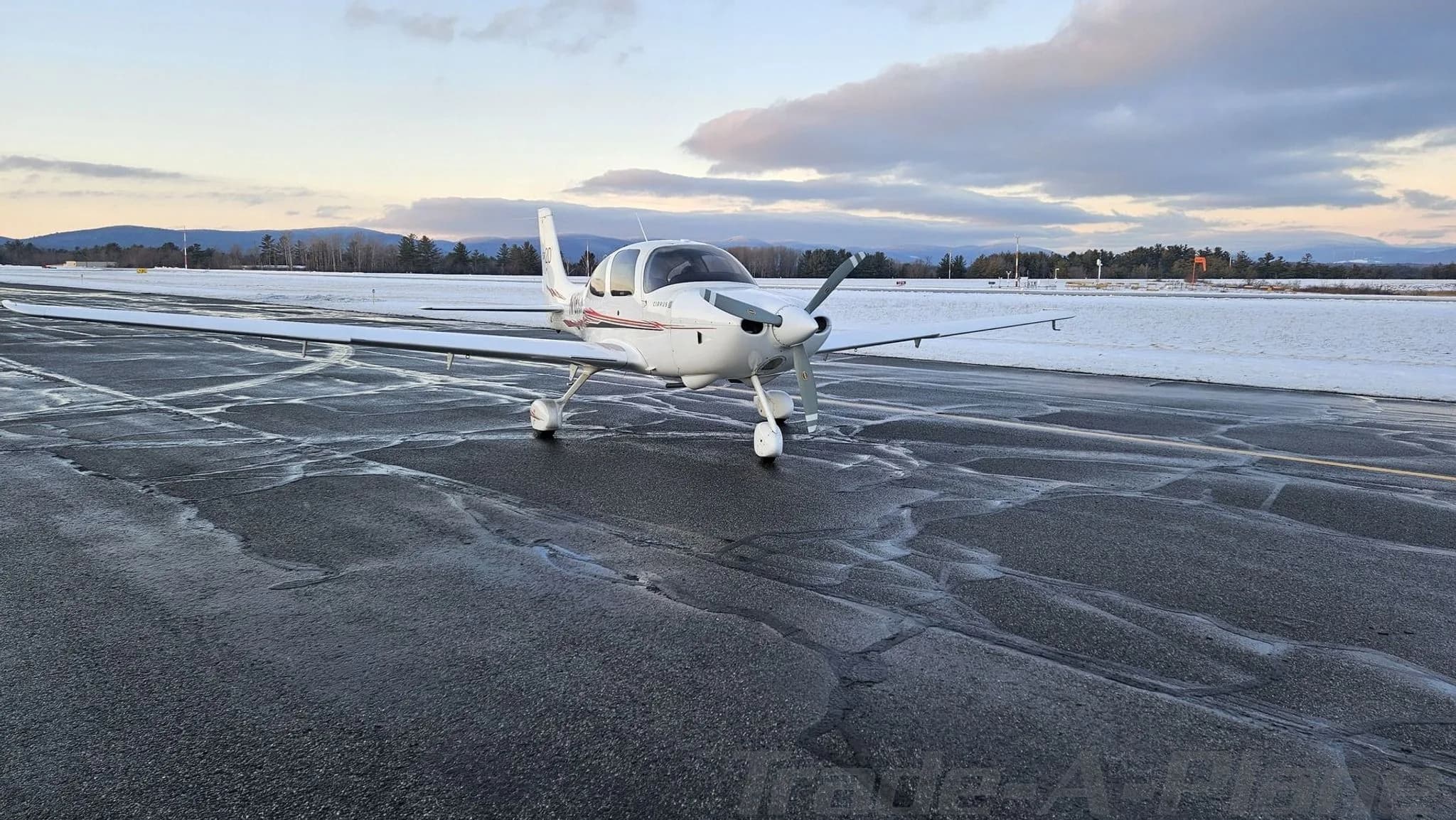 Front view of a white Cirrus SR20 aircraft with red and black stripes on a wet tarmac.