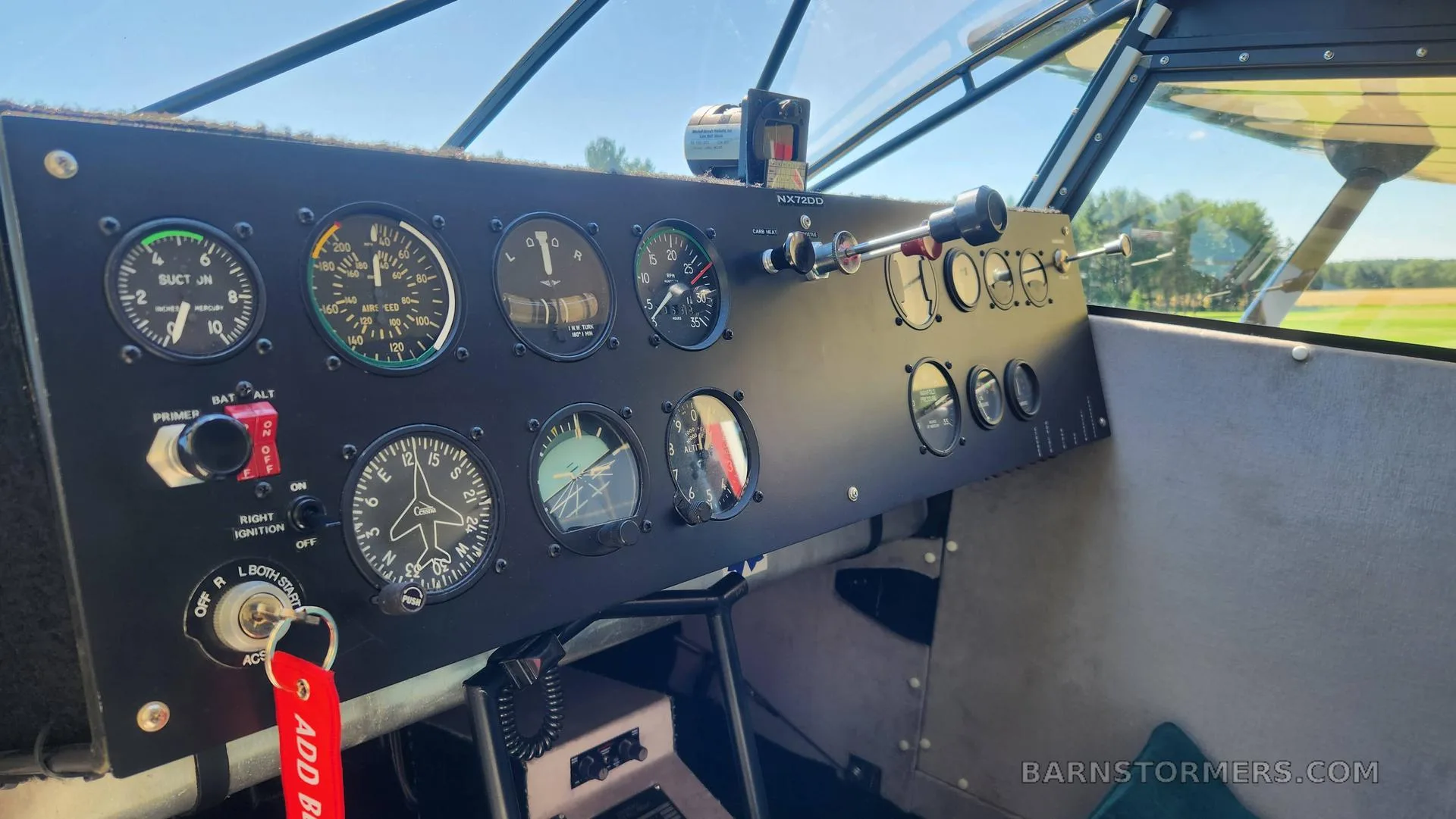The black instrument panel of a small aircraft, showing various gauges and controls, with a view of a field outside.