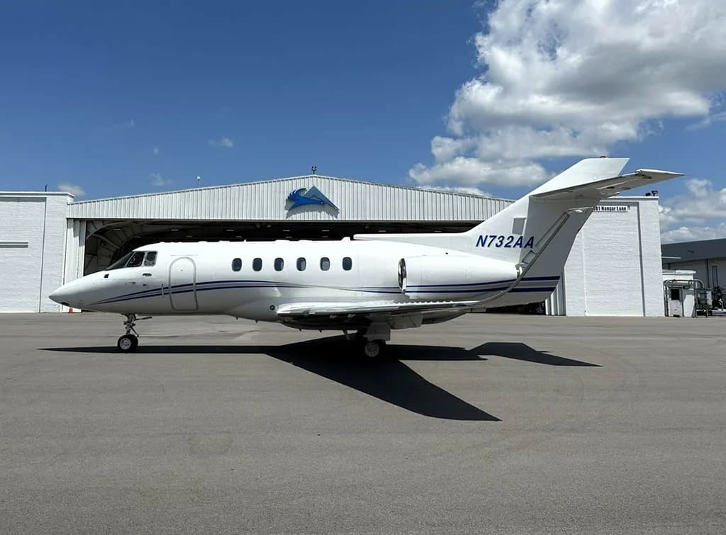 A side view of a white business jet, N732AA, with blue stripes parked on an tarmac in front of a white hangar.