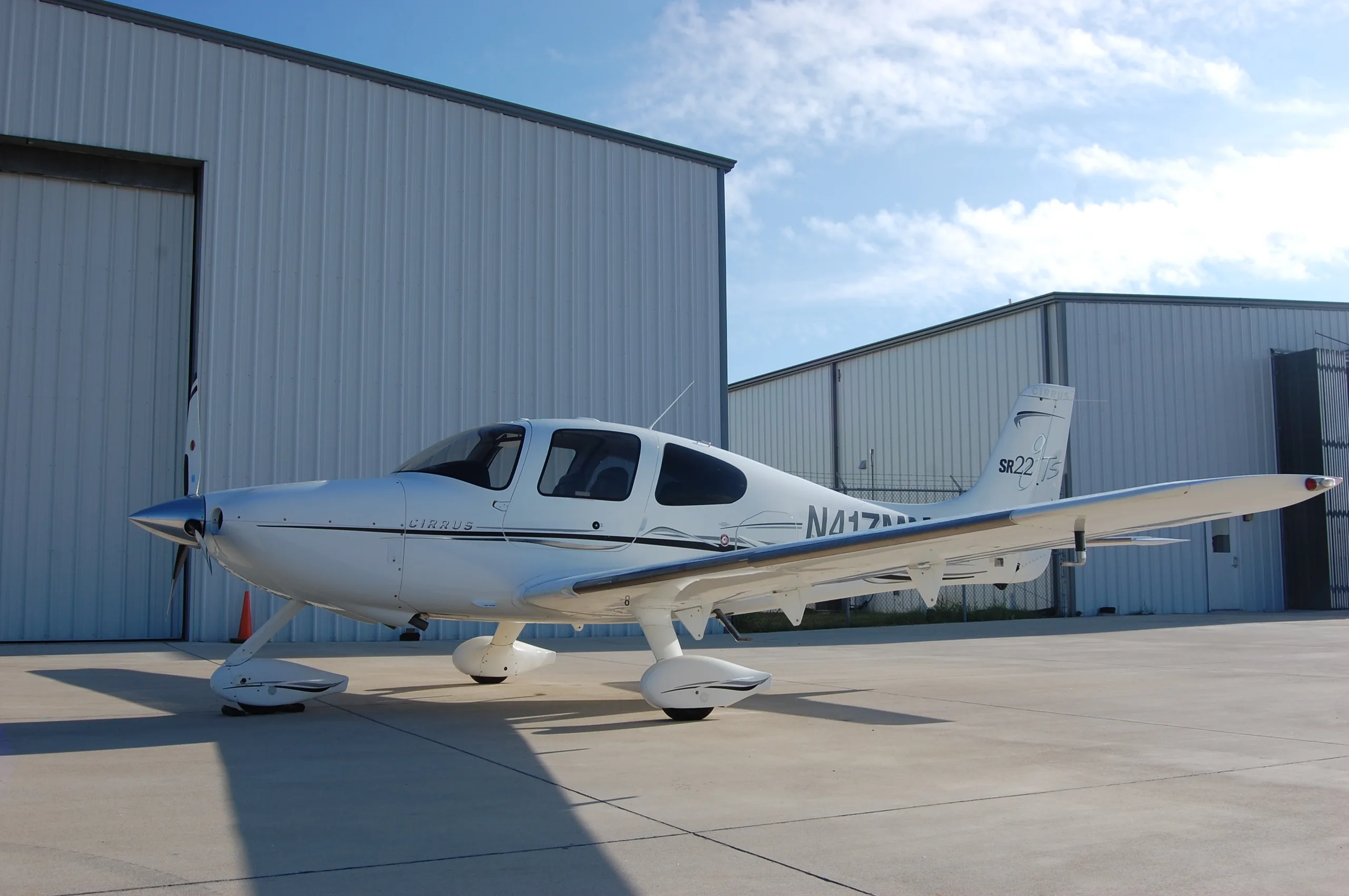 A white Cirrus SR22 aircraft, N417MM, parked on an airfield with hangars under a blue sky.