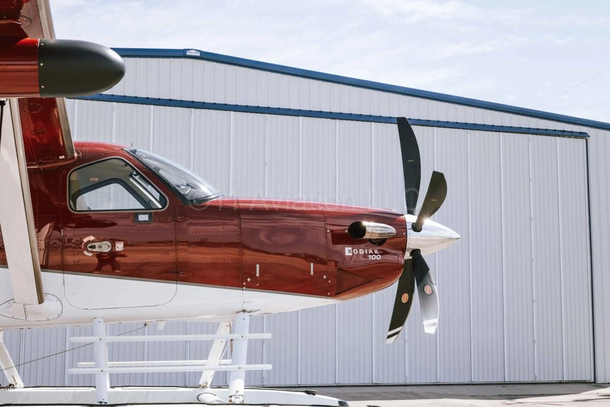 A close-up view of the front-right side of a red and white Kodiak 100 aircraft with a propeller, parked on an airfield.