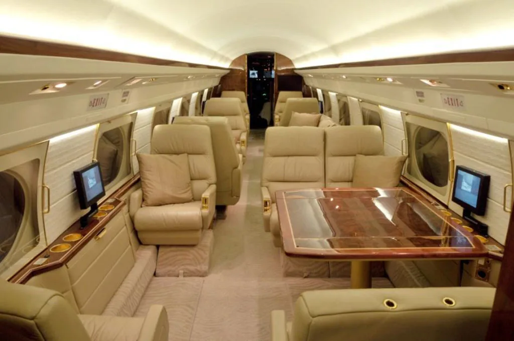 A wider view of the private jet's interior cabin, featuring multiple beige leather seats, a large wooden table, and a sofa.