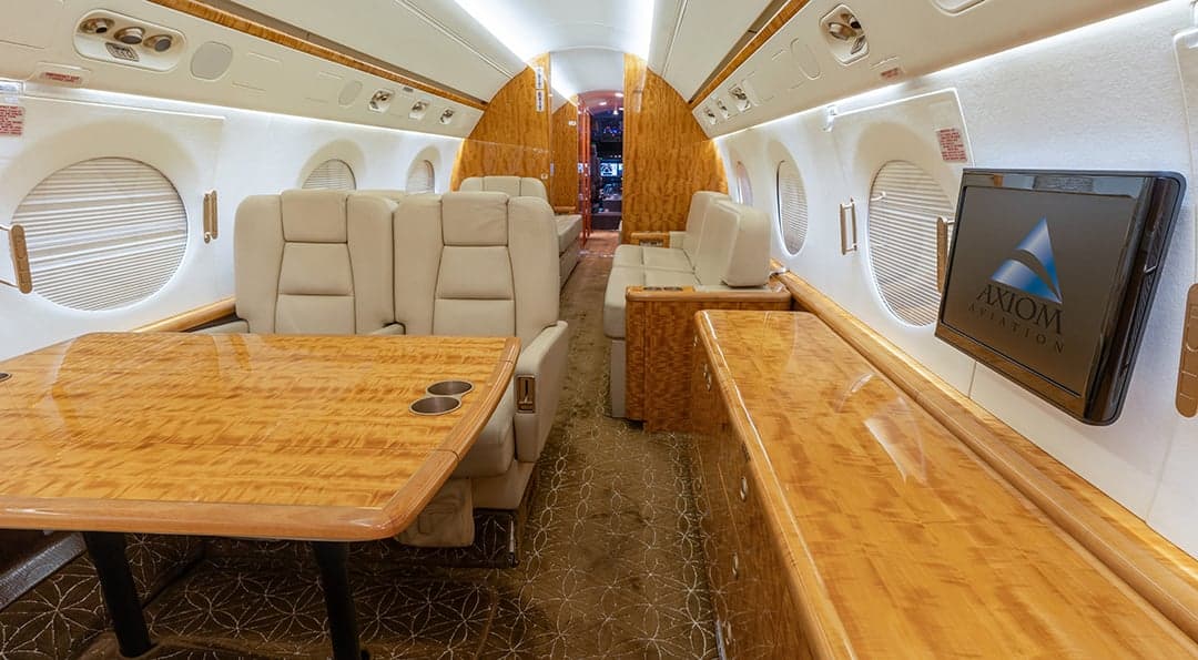 The aircraft's interior features a table, cream-colored leather seats, and a monitor displaying the Axiom Aviation logo.