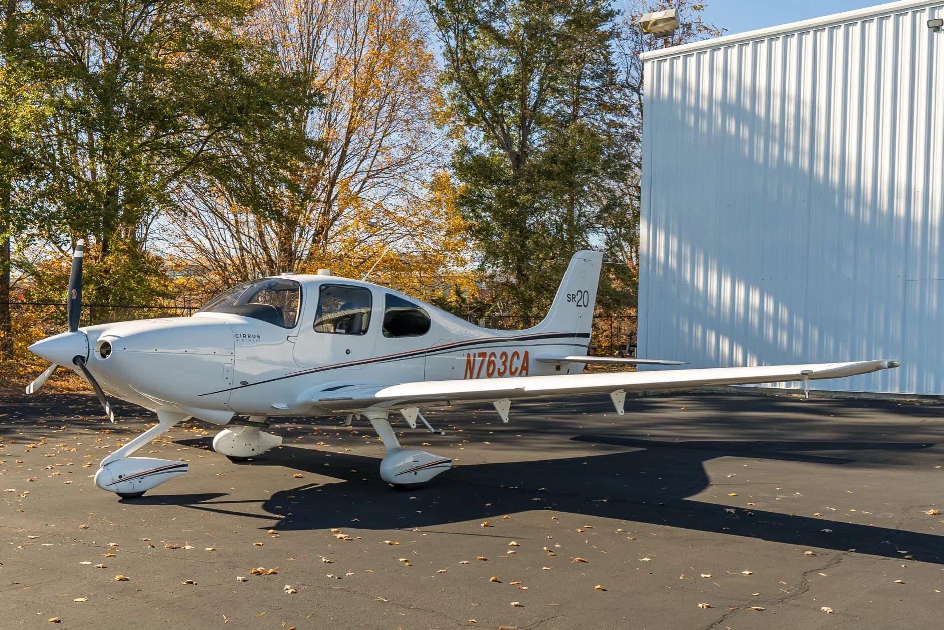 A white Cirrus SR20 aircraft with orange and black stripes is parked on an asphalt tarmac with autumn trees in the background.