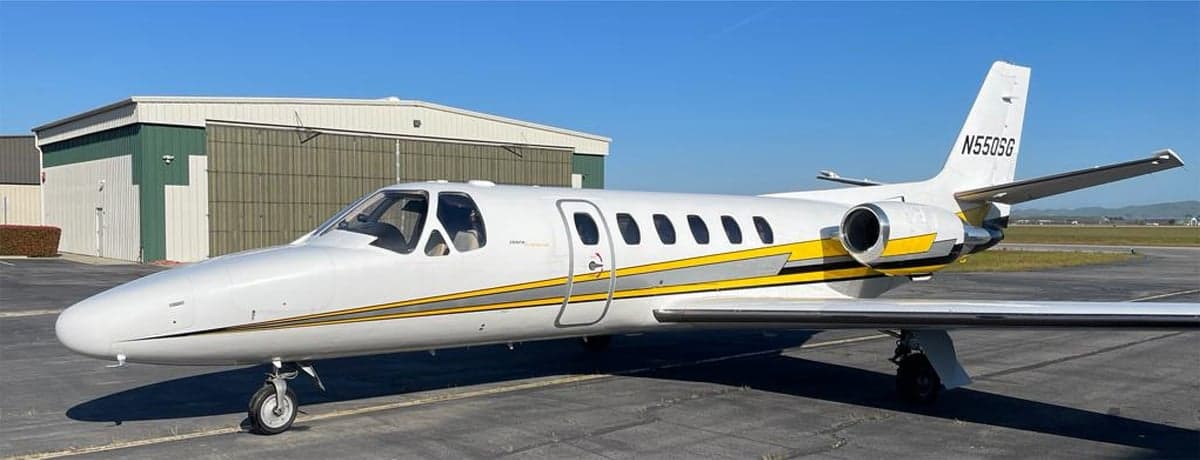 A Cessna Citation CJ3 with a white and yellow paint scheme is parked on the tarmac in front of a hangar.