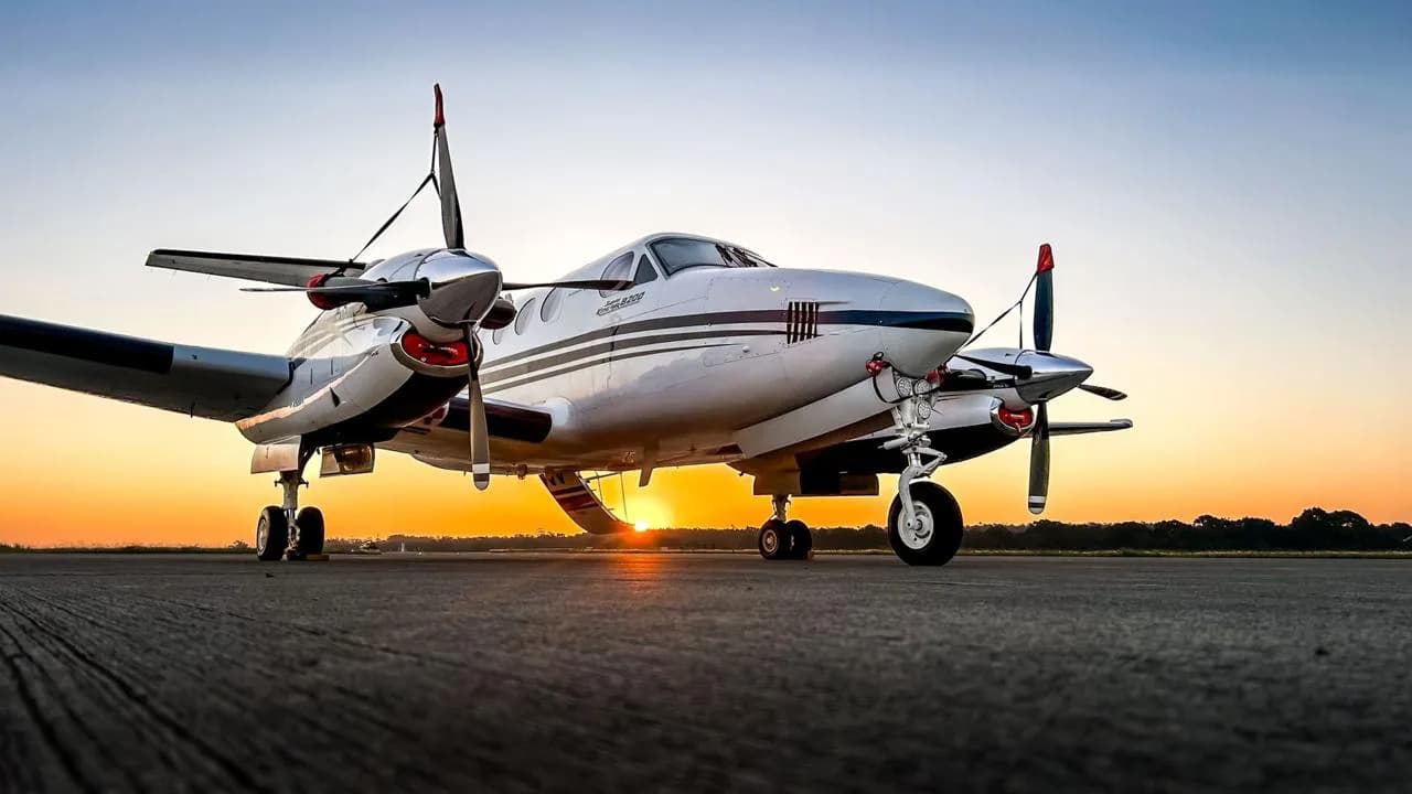 A white and black Beechcraft King Air aircraft is parked on an airfield at sunset, viewed from a low angle.