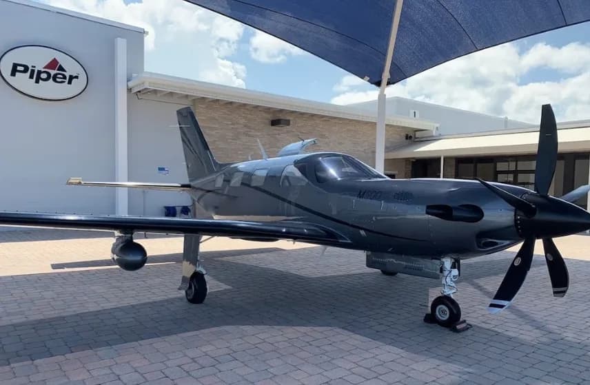 A dark grey Piper M600 SLS aircraft is parked on a paved area next to a building with a Piper logo.