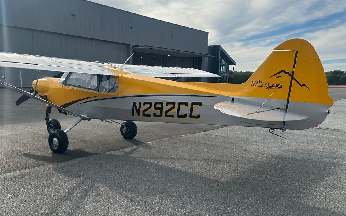 A yellow and gray NX Cub aircraft, N292CC, parked on an asphalt tarmac next to a modern hangar.