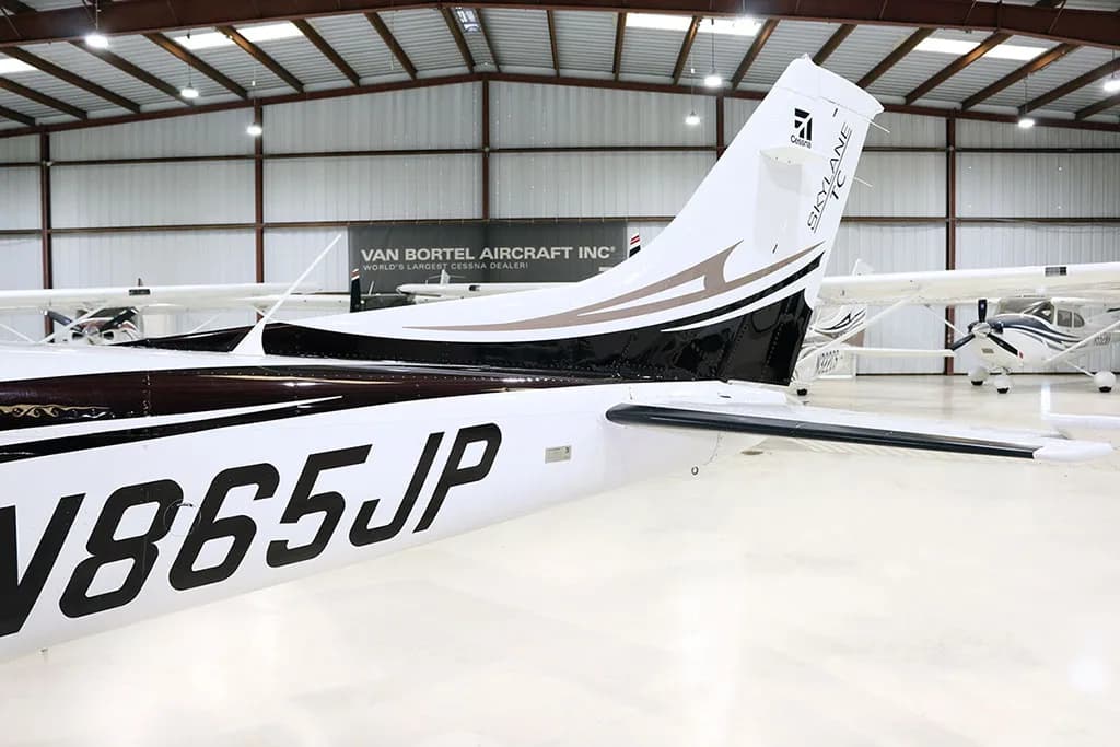 The tail section of a white and black Cessna Skylane N865JP with brown accents is shown in a hangar.