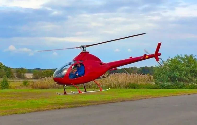 A red RotorWay Exec helicopter with a pilot and passenger hovers low over a grassy field.