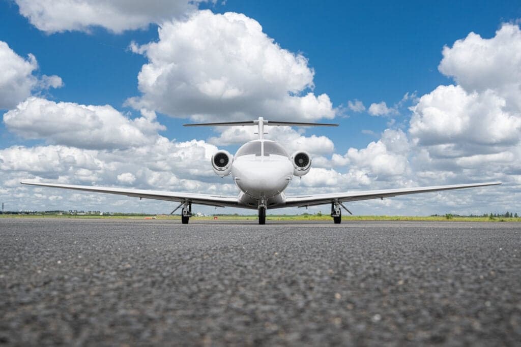 A white Cessna Citation CJ3 (D-IALI) private jet seen head-on from a low angle on an asphalt runway under a blue sky with white clouds.