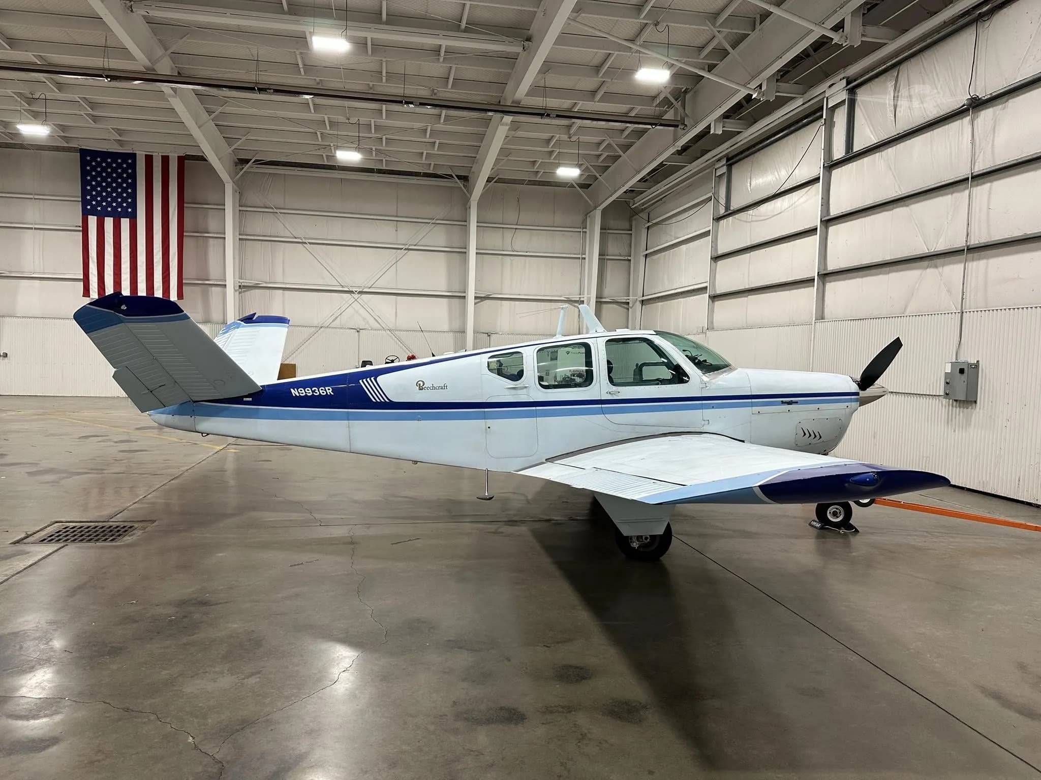 A white and blue Beechcraft Bonanza N9936R parked in a hangar with an American flag on the wall.