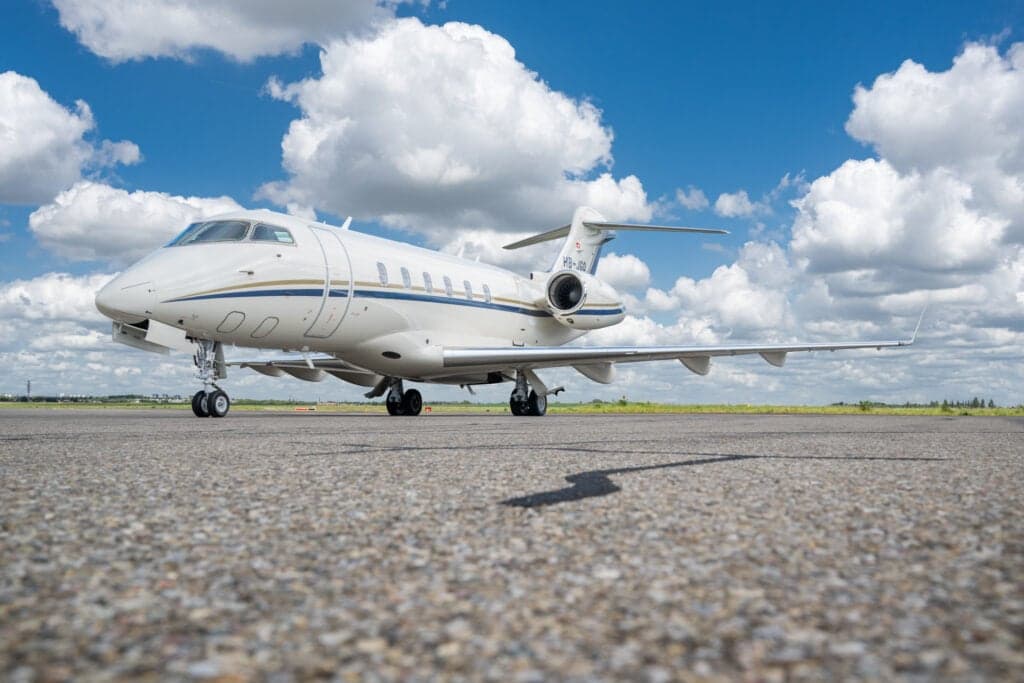 A Bombardier Challenger 350 aircraft with a white and gold paint scheme is parked on the tarmac under a blue sky with scattered clouds.