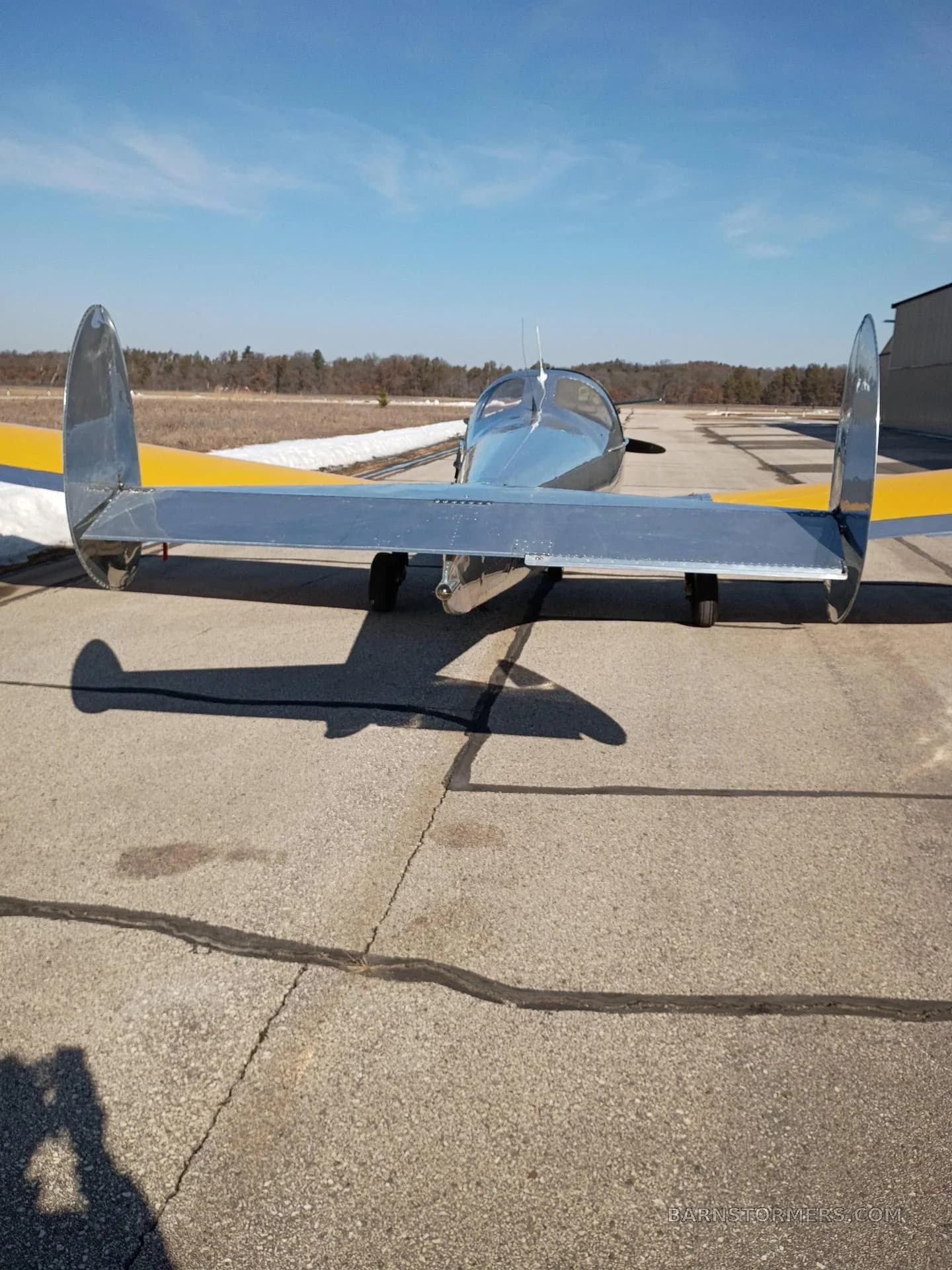 A rear view of a silver aircraft with yellow wings and tail, parked on an airfield.