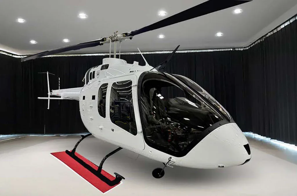 A white Bell 505 helicopter with black rotors is displayed in a brightly lit hangar.