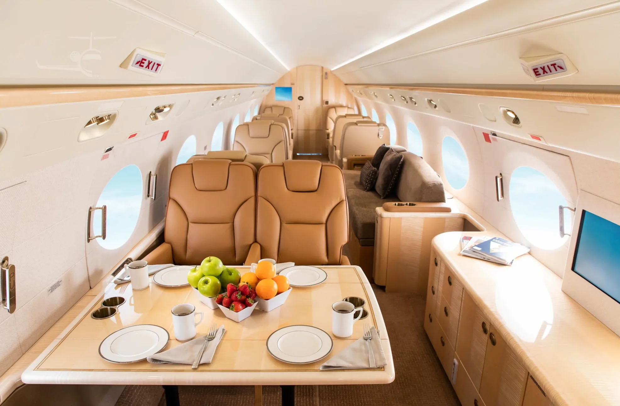 A luxurious private jet cabin with tan leather seats, a dining table set with fruit, and large windows.