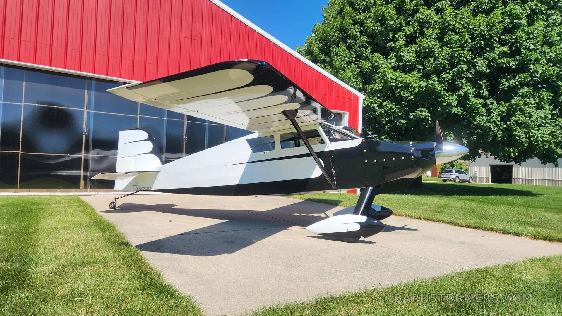 A black and white single-engine aircraft with tail number NXZ2DD is parked on concrete, viewed from the front-left, next to a red hangar.