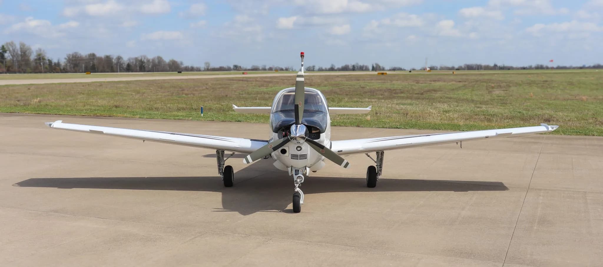 A head-on view of a white Beechcraft Bonanza G36 aircraft with a black nose, parked on an airfield with a grassy background.