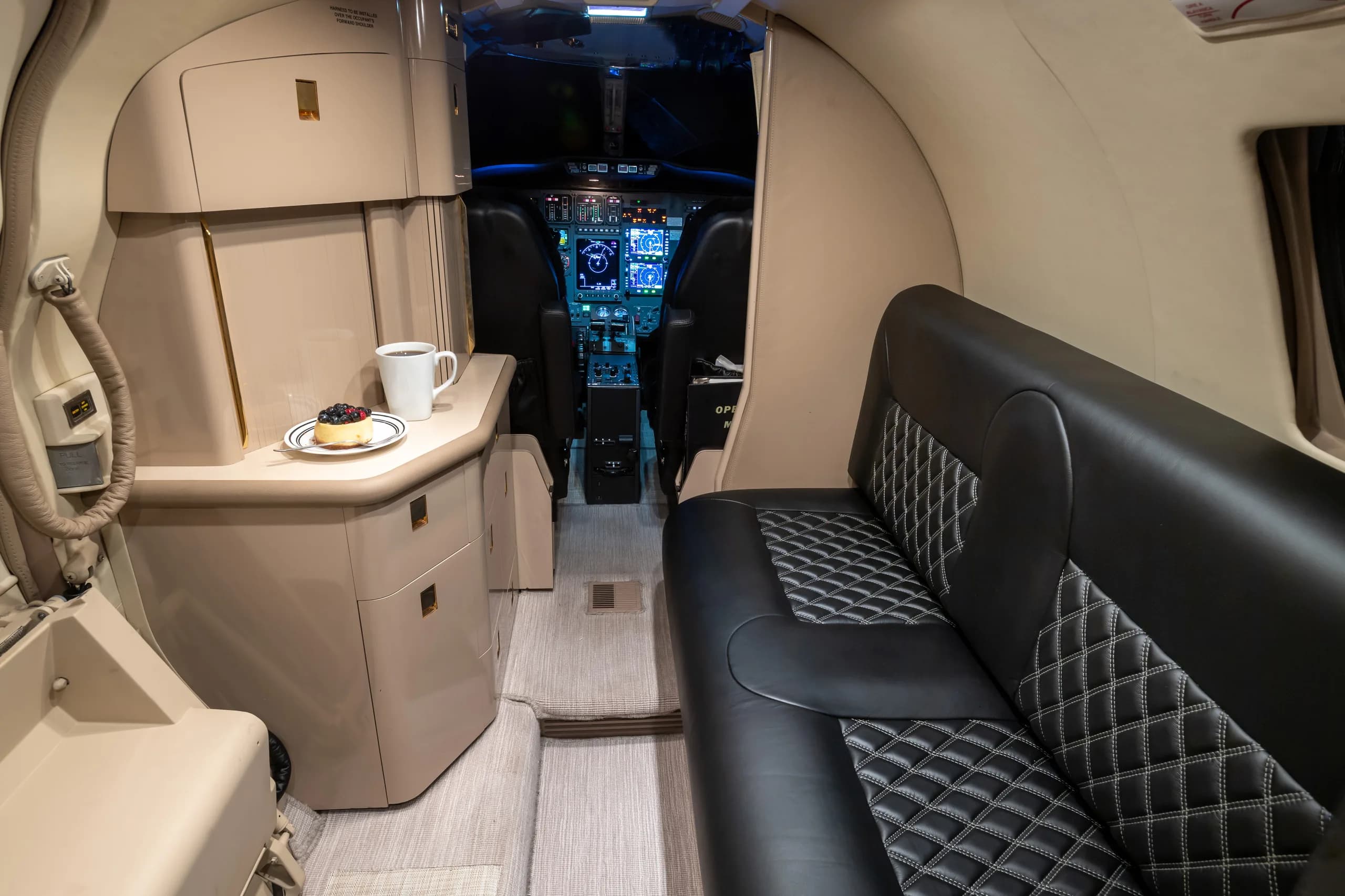 View from the rear of the business jet cabin, showing a side couch, a galley area, and a clear view into the modern cockpit.