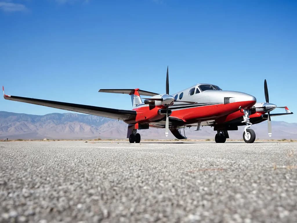 Low-angle view of a red and silver Beechcraft King Air N350FC on an airfield with mountains in the background.