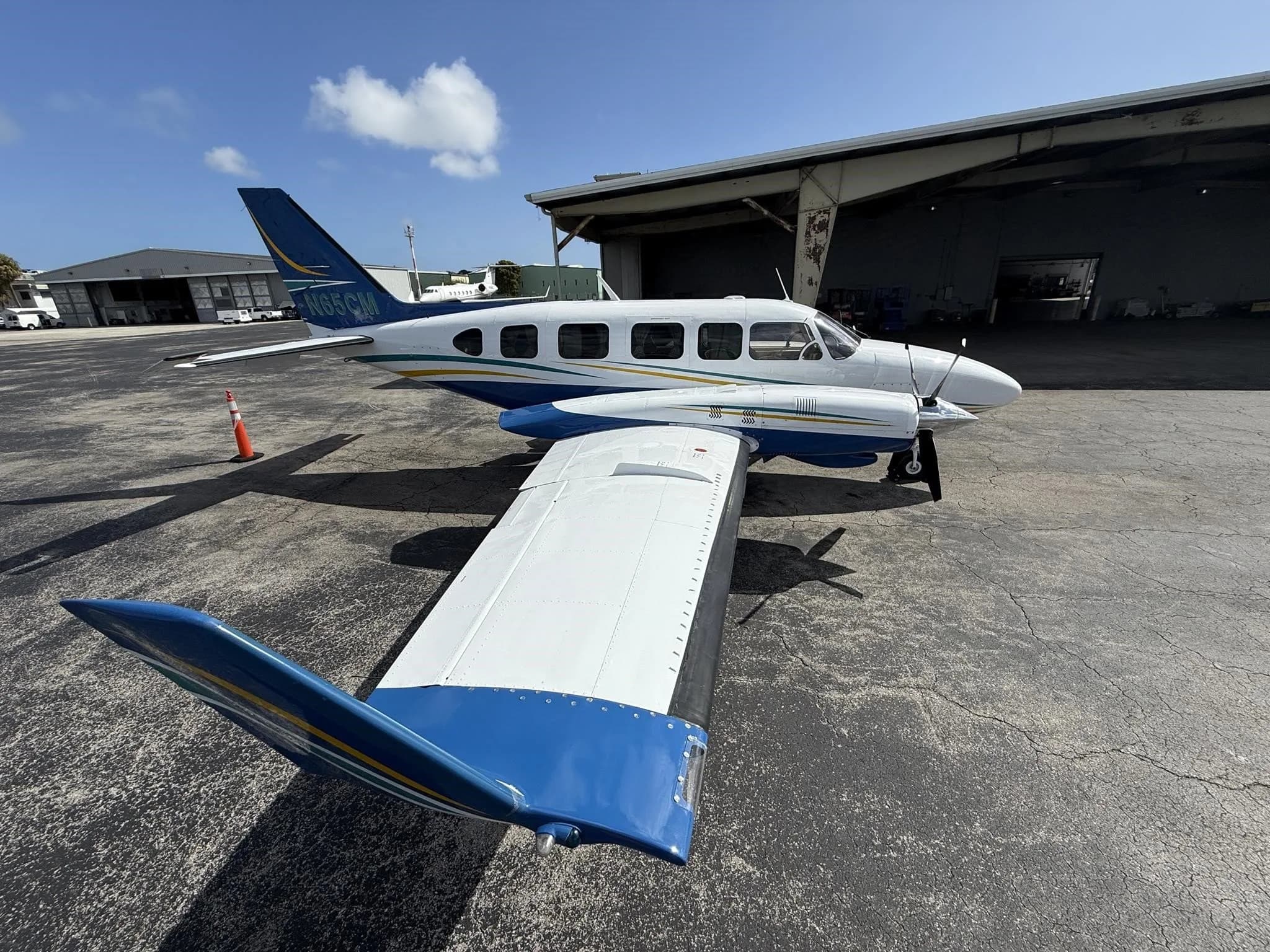 A rear-side view of a white and blue twin-engine propeller aircraft with tail number N65CM, parked next to a hangar.