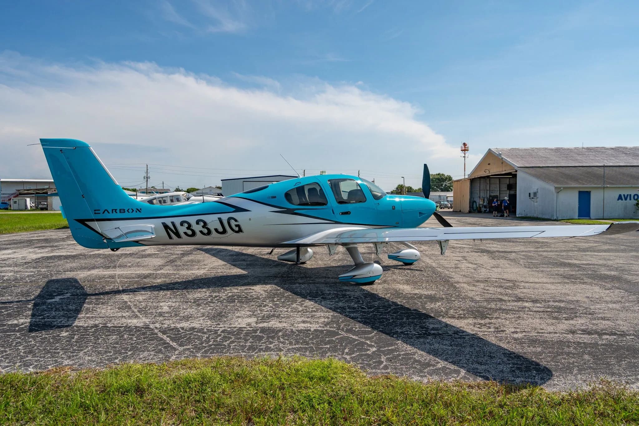 A blue and silver Carbon aircraft (N33JG) parked on an asphalt ramp with a white hangar building in the background.