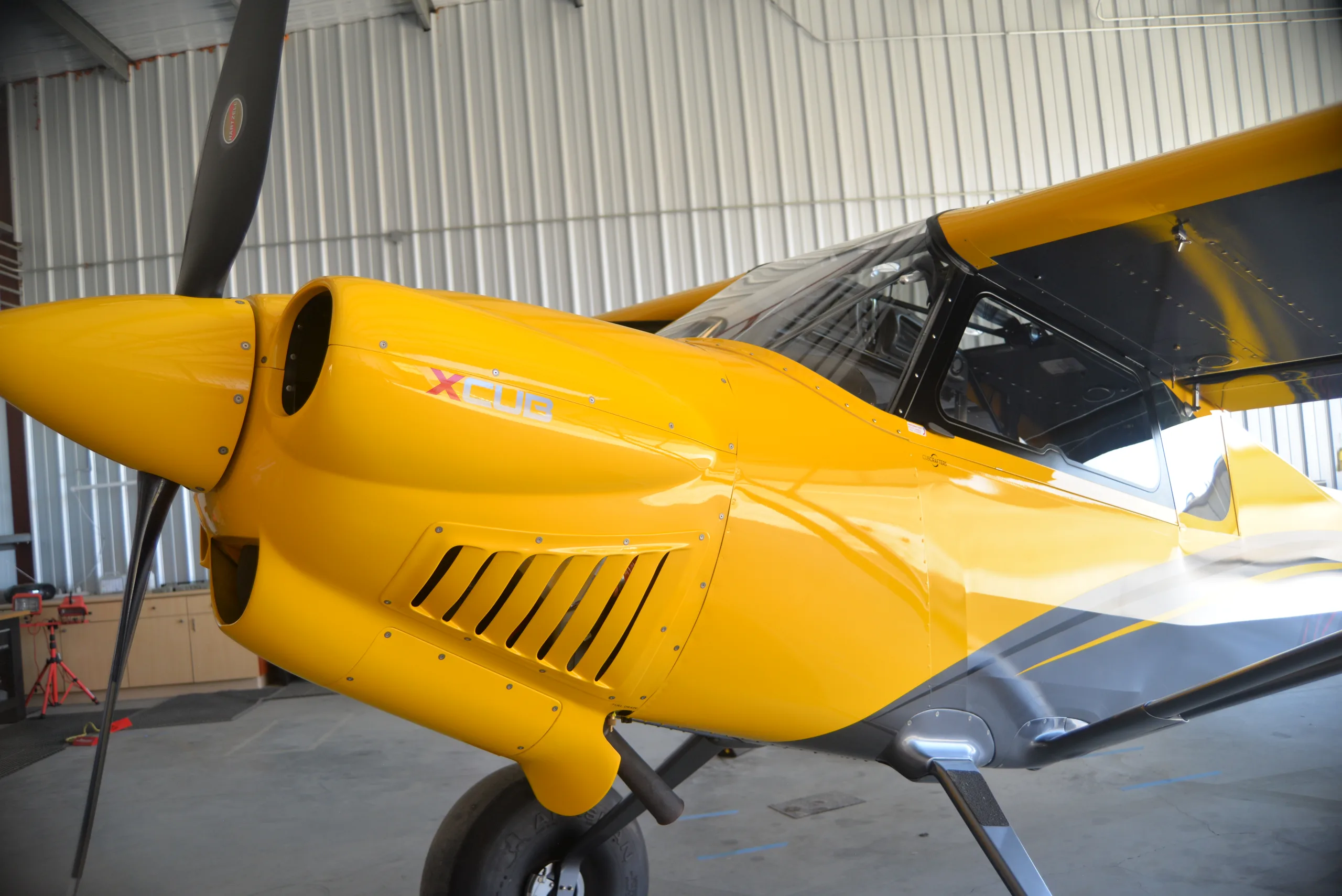 Close-up of the cowling of a yellow CubCrafters XCub, registration N27MB, inside a hangar.