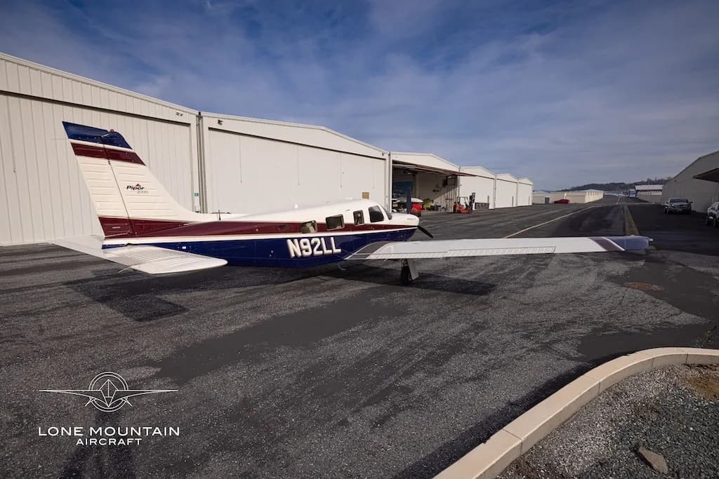 A rear three-quarter view of the Piper Saratoga II TC aircraft (N92LL) with blue and white paint scheme parked on an asphalt apron next to hangars.
