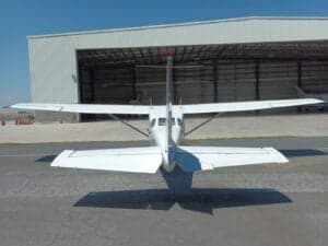 Rear view of a white Cessna aircraft with its tail number obscured, parked in front of a hangar.