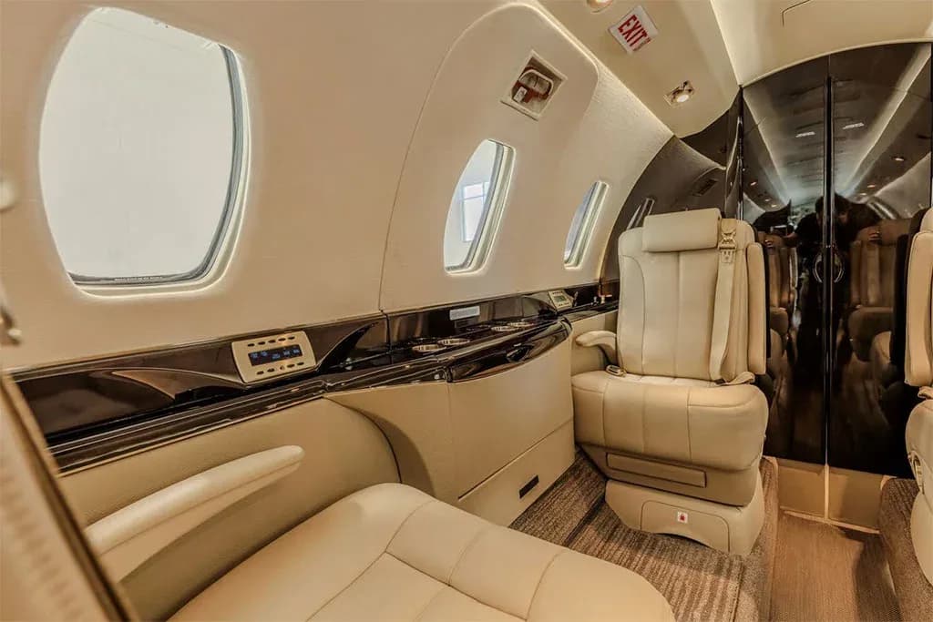 A close-up view of the private jet's interior cabin, showing a beige leather seat, side panel controls, and an "EXIT" sign.