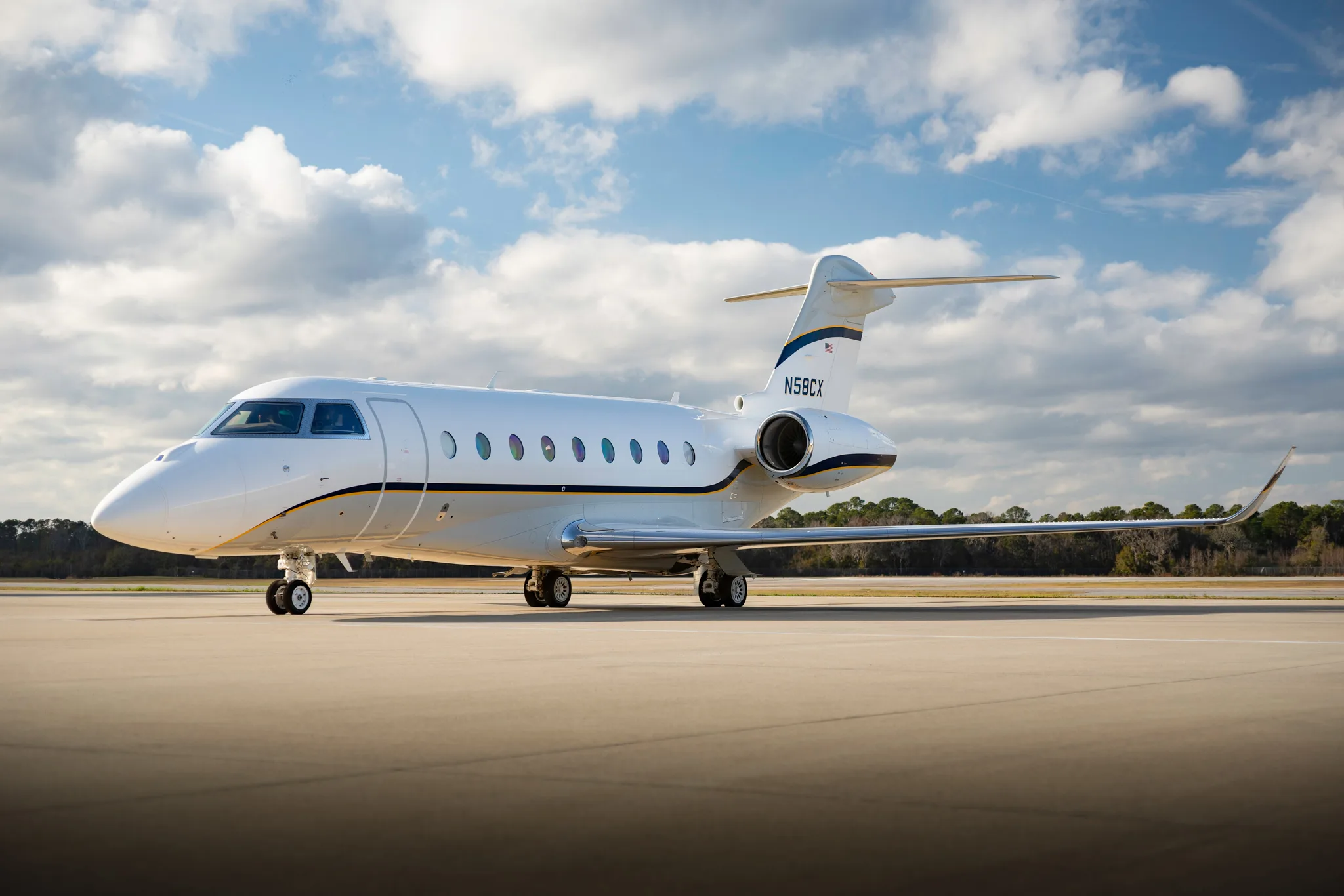 A white business jet with blue and gold stripes, registration N58CX, is parked on a tarmac under a partly cloudy blue sky.