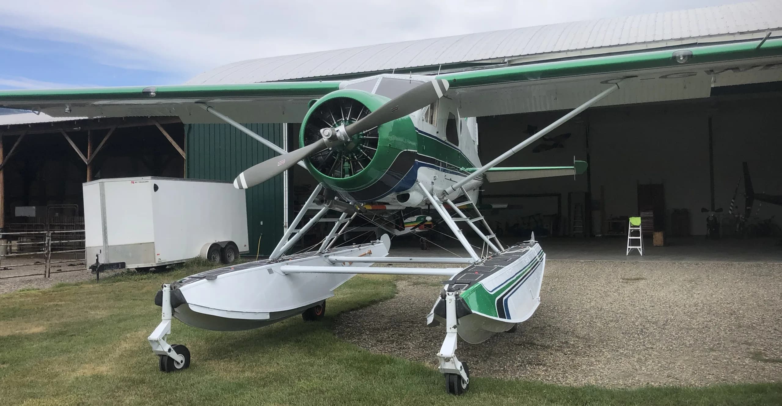 A green, white, and blue single-engine floatplane with retractable landing gear on its floats, parked on gravel in front of a hangar.