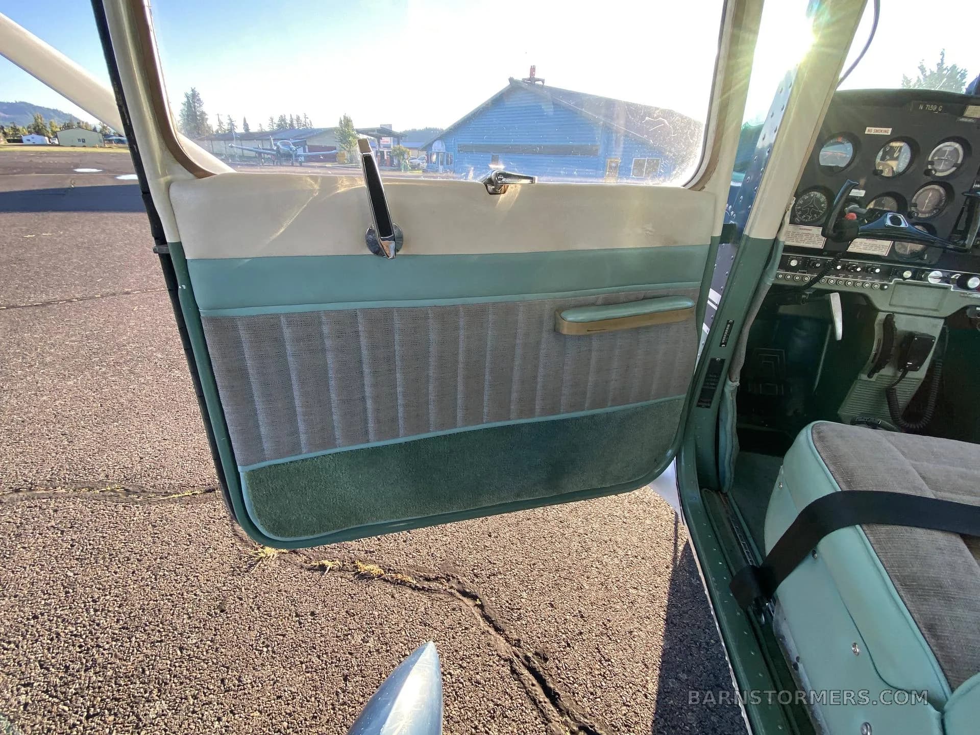 The open door of a light aircraft reveals a green and beige interior with a partial view of the cockpit instruments.