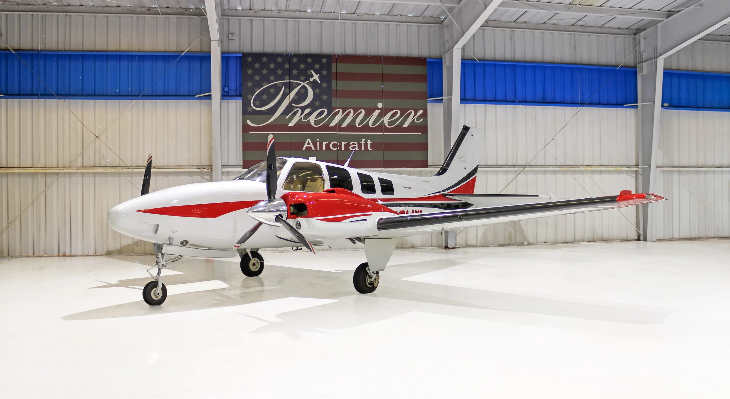 A white and red twin-engine propeller aircraft, a Beechcraft Baron, parked in a hangar.