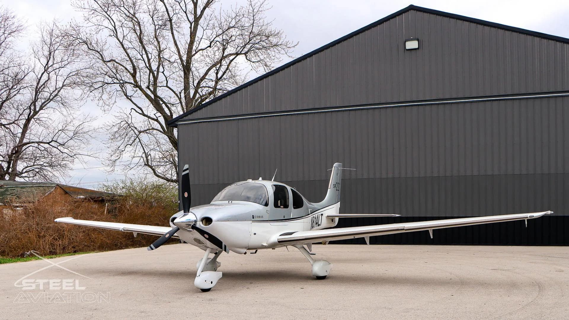 A three-quarter front view of a white and silver Cirrus SR22 aircraft (N814LT) parked on an apron next to a dark grey hangar.