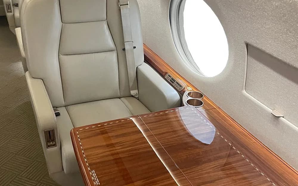 A close-up of a light beige leather cabin seat next to a large window, with a polished wooden side table featuring cup holders.