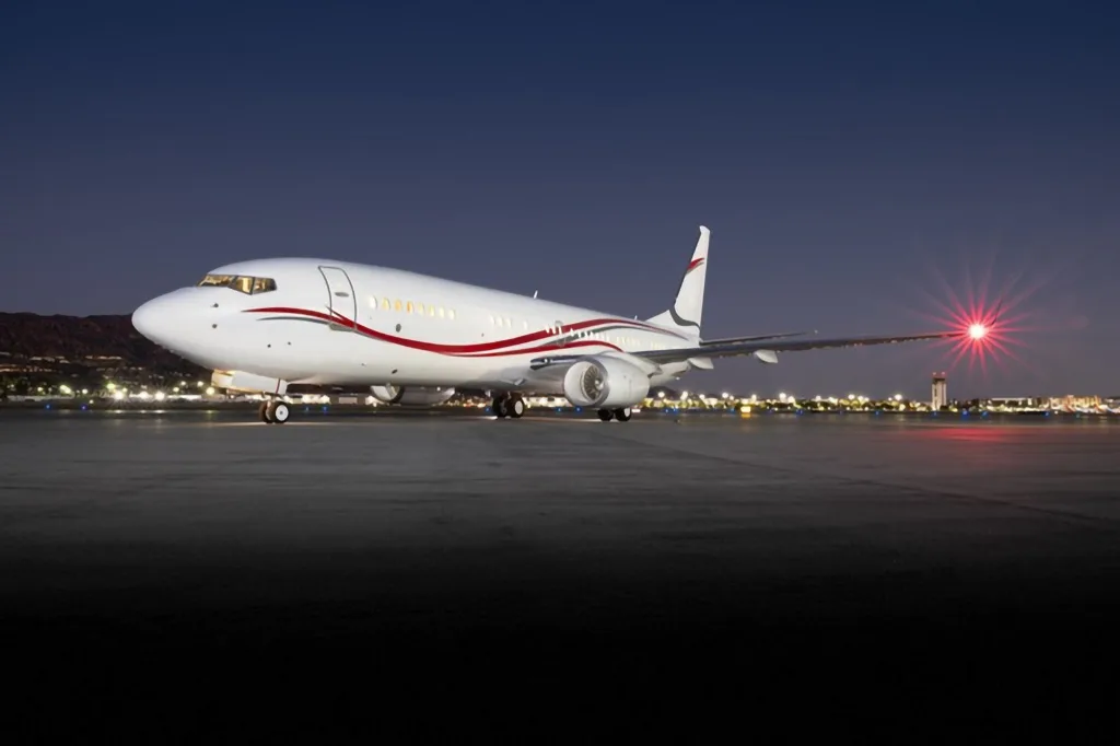 A white Boeing 737 BBJ with red and black stripes on the fuselage, parked on a tarmac at night with city lights in the background.