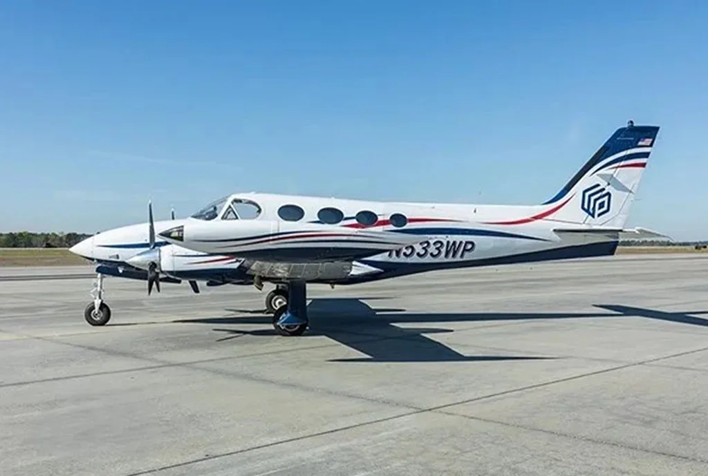 A side view of a white twin-engine propeller aircraft with blue and red stripes, parked on an airfield.